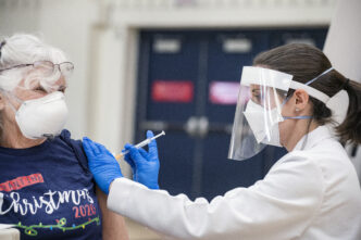 Impacto Covid-19: Mujer recibiendo vacuna contra el Covid-19 aplicada por personal médico con equipo de protección. Medidas comerciales y sanitarias.
