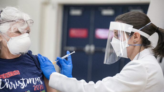 Impacto Covid-19: Mujer recibiendo vacuna contra el Covid-19 aplicada por personal médico con equipo de protección. Medidas comerciales y sanitarias.