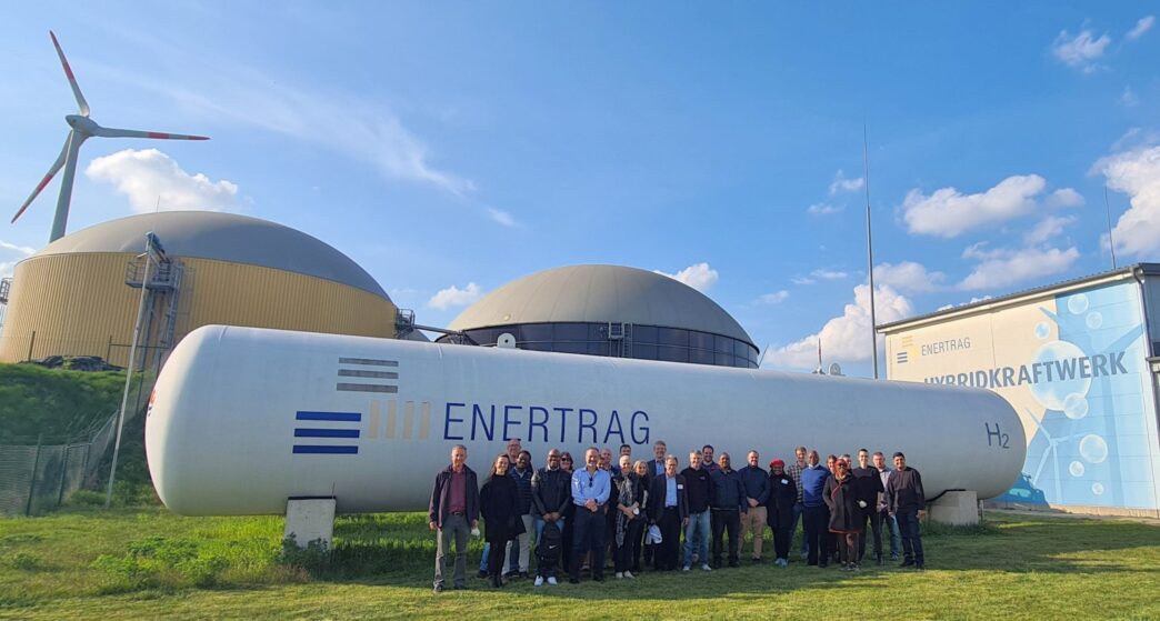 Grupo frente a tanque de hidrógeno ENERTRAG en Alemania, con aerogenerador y planta híbrida. Innovación en infraestructura verde y redes de hidrógeno.