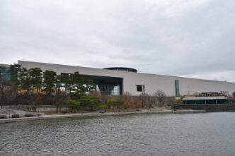 Fachada del Museo Nacional de Literatura Coreana en Seúl, con un lago en primer plano y árboles alrededor. Arquitectura moderna y cielo nublado.