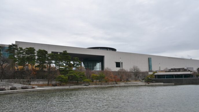 Fachada del Museo Nacional de Literatura Coreana en Seúl, con un lago en primer plano y árboles alrededor. Arquitectura moderna y cielo nublado.