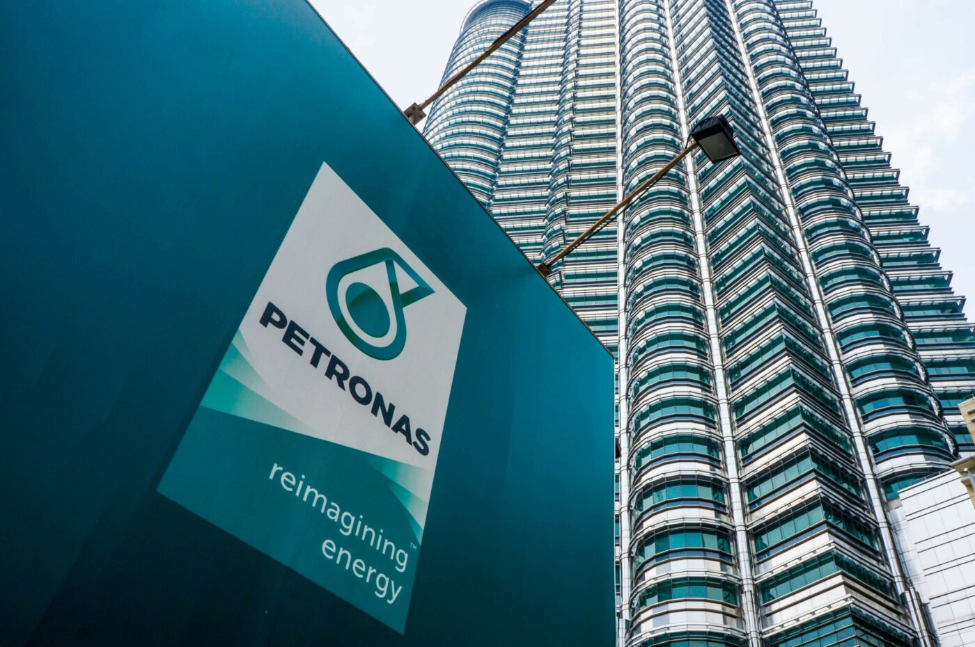 Primer plano del logo de PETRONAS frente a las torres gemelas, destacando innovaciones sostenibles en infraestructura petrolera. Re-imaginando energía.