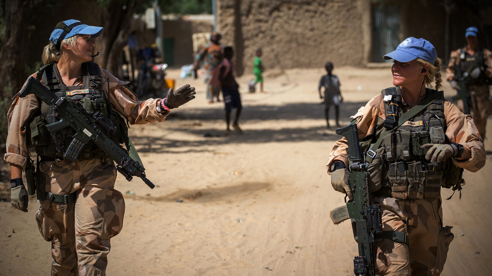 Dos militares de la ONU patrullan una calle polvorienta. Los soldados, con uniforme y armas, interactúan en un entorno urbano africano.