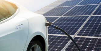 Carga EV con energía solar: coche eléctrico blanco cargando frente a paneles solares. Integración solar y carga EV para real estate sostenible.