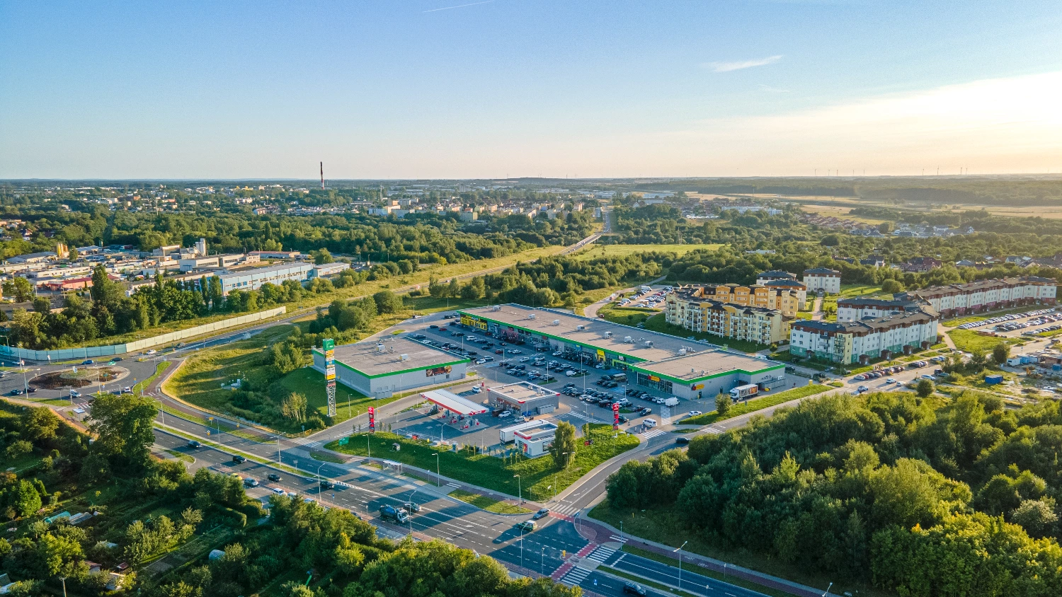 Vista aérea de inversión sostenible en real estate polaco: centro comercial con parking, gasolinera y edificios residenciales en un entorno verde.