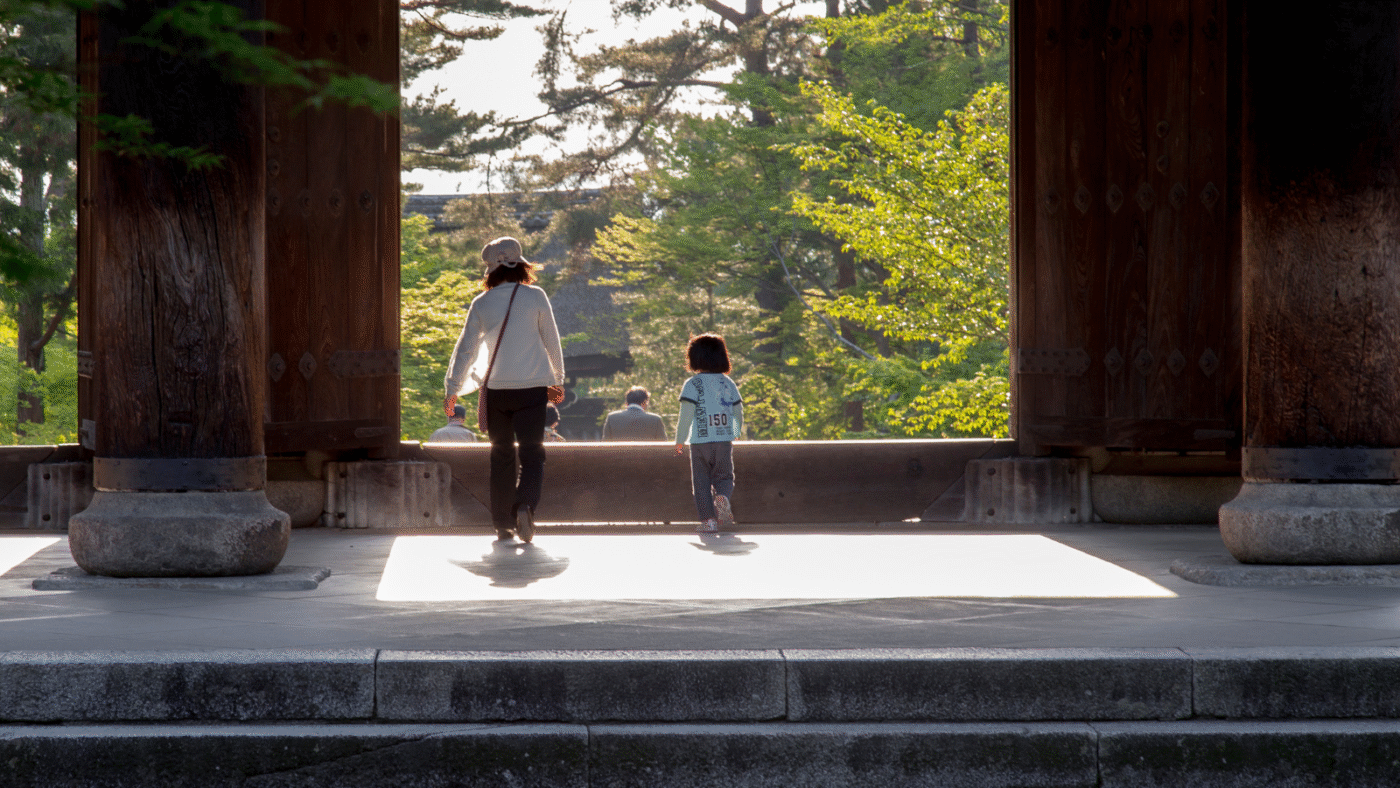 Arquitectura tradicional en [city], Japón. Una mujer y un niño caminan a través de una puerta de madera hacia un jardín iluminado por el sol.