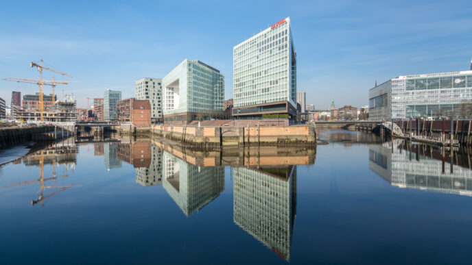 Arquitectura moderna en Bokarina reflejada en el agua, con edificios de diseño y un horizonte urbano transformado. Detalles de la construcción y cielo azul.