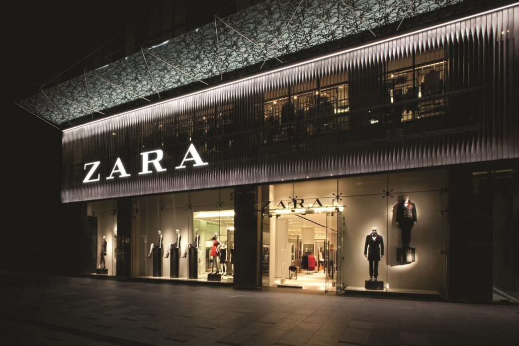 Fachada iluminada de Zara, un ejemplo de retail tech en el lujo arquitectónico. Maniquíes exhiben moda actual tras los grandes ventanales.