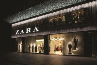 Fachada iluminada de Zara, un ejemplo de retail tech en el lujo arquitectónico. Maniquíes exhiben moda actual tras los grandes ventanales.