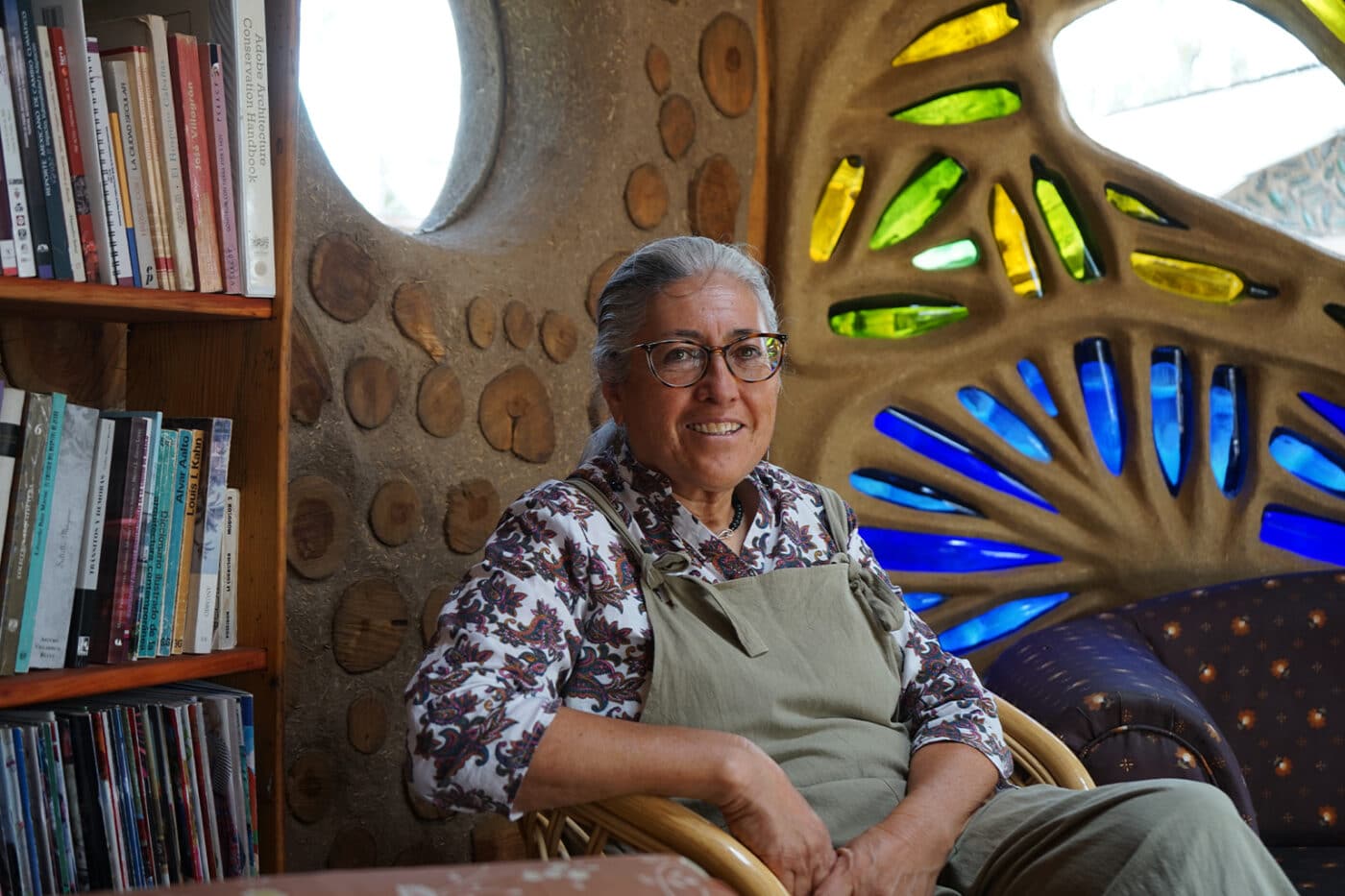 Mujer sonriente en ambiente de bioconstrucción, líder en México. Estantería con libros y pared con detalles de madera y vidrio colorido.