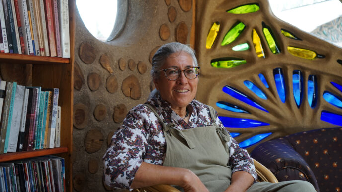 Mujer sonriente en ambiente de bioconstrucción, líder en México. Estantería con libros y pared con detalles de madera y vidrio colorido.