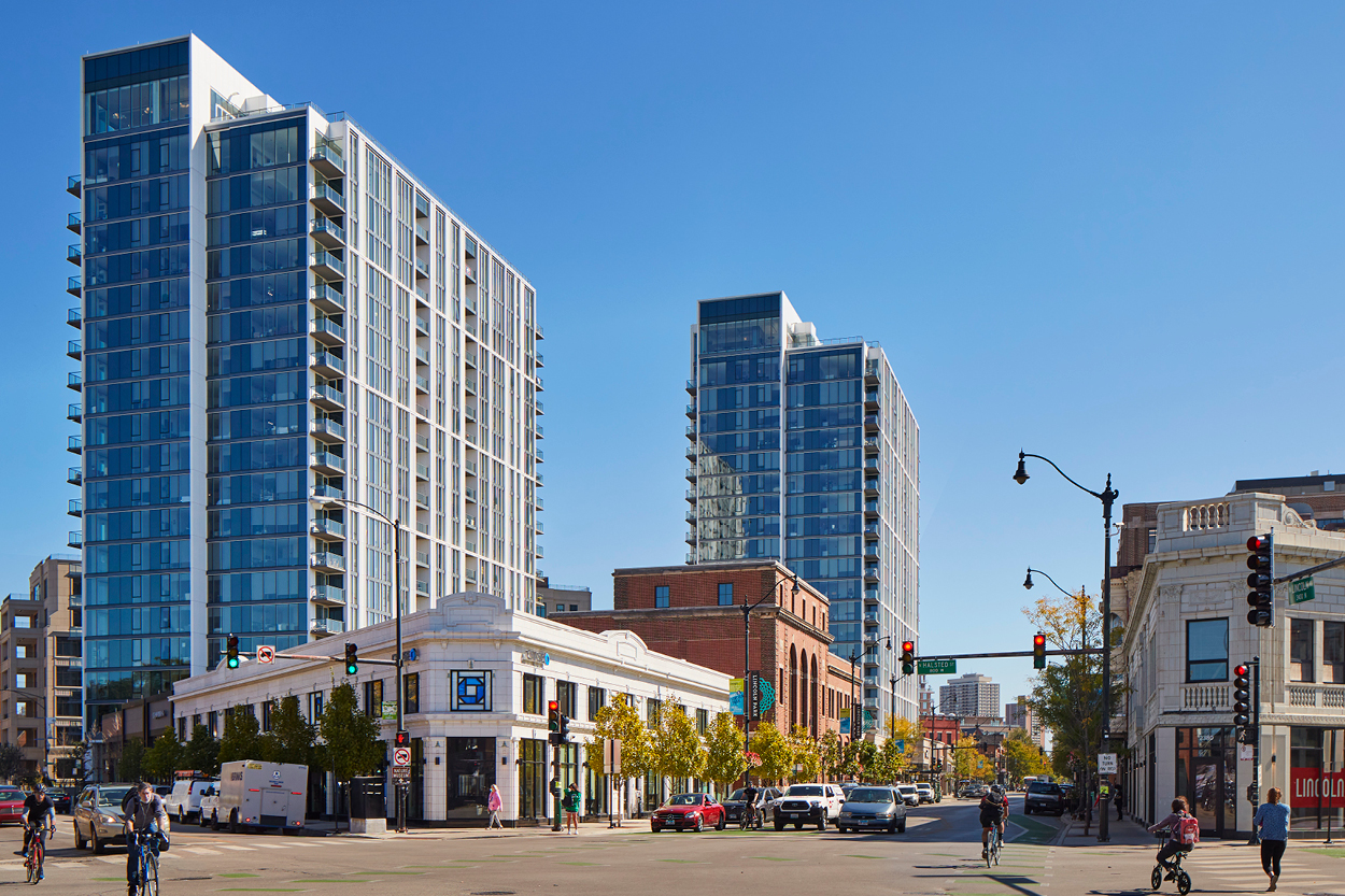 Desarrollo BTR Naperville por Lincoln Property Co.: Dos torres modernas dominan el paisaje urbano, con tráfico y peatones en una calle arbolada.