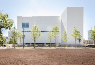 Vista exterior del Dovetail Building. Multiplex asegura contrato millonario. Fachada blanca, árboles jóvenes y explanada en primer plano. Diseño arquitectónico.
