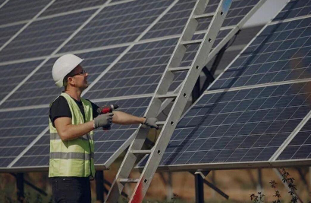 Instalador con casco ajustando paneles solares para construcción eficiente energéticamente. Escalera y herramientas visibles. NJBPU impulsa la energía solar.