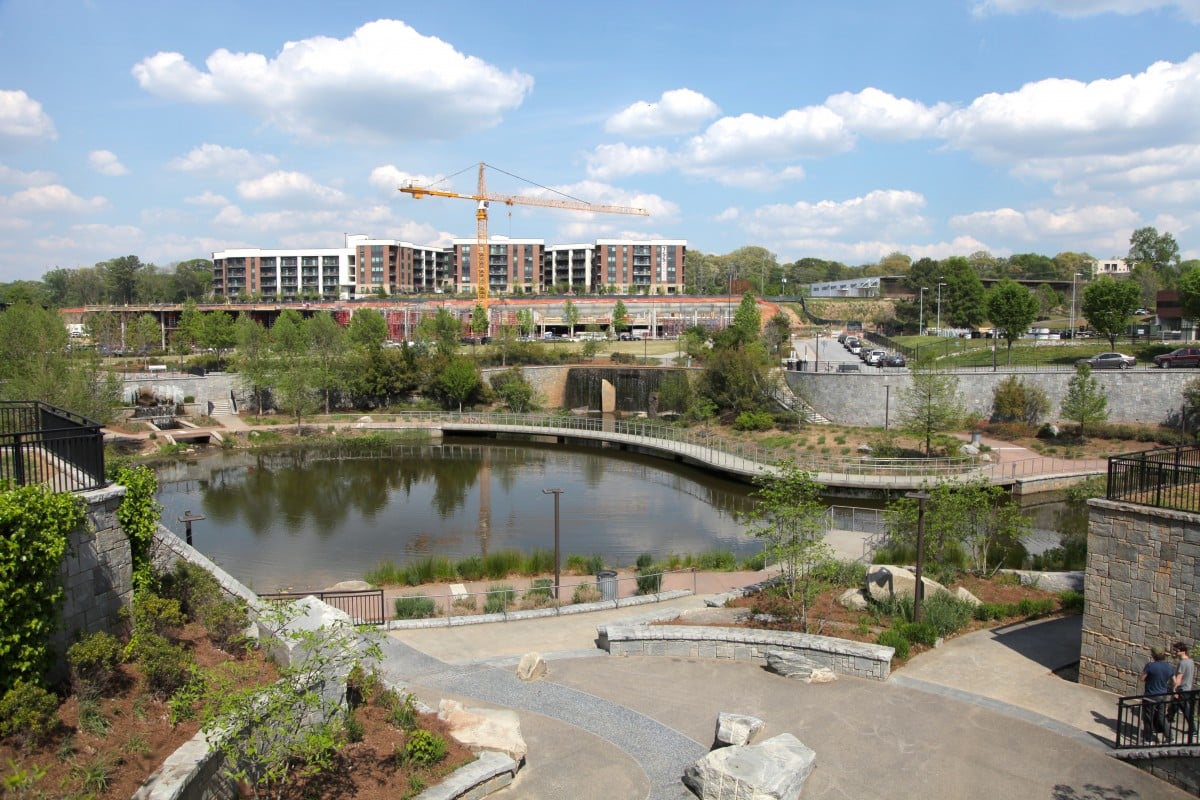Vista panorámica de un parque urbano con un lago, puente peatonal, edificios residenciales y una grúa, resaltando el desarrollo de ciudades.