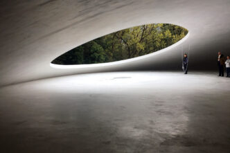 Interior arquitectónico con estructura de piedra natural y abertura ovalada que enmarca vegetación. Personas en el espacio resaltan la escala, 125c