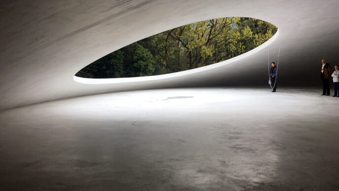 Interior arquitectónico con estructura de piedra natural y abertura ovalada que enmarca vegetación. Personas en el espacio resaltan la escala, 125c