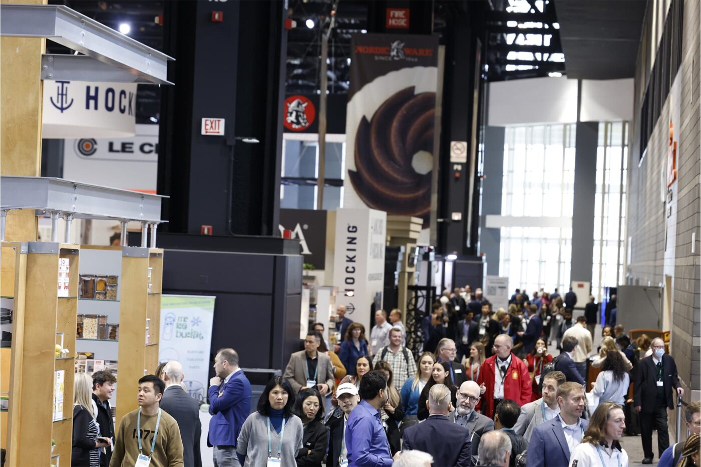 Multitud en feria comercial. Stands de exhibición, incluyendo 'Hocking' y 'Nordic Ware'. Ambiente concurrido, enfoque en la variedad de productos.