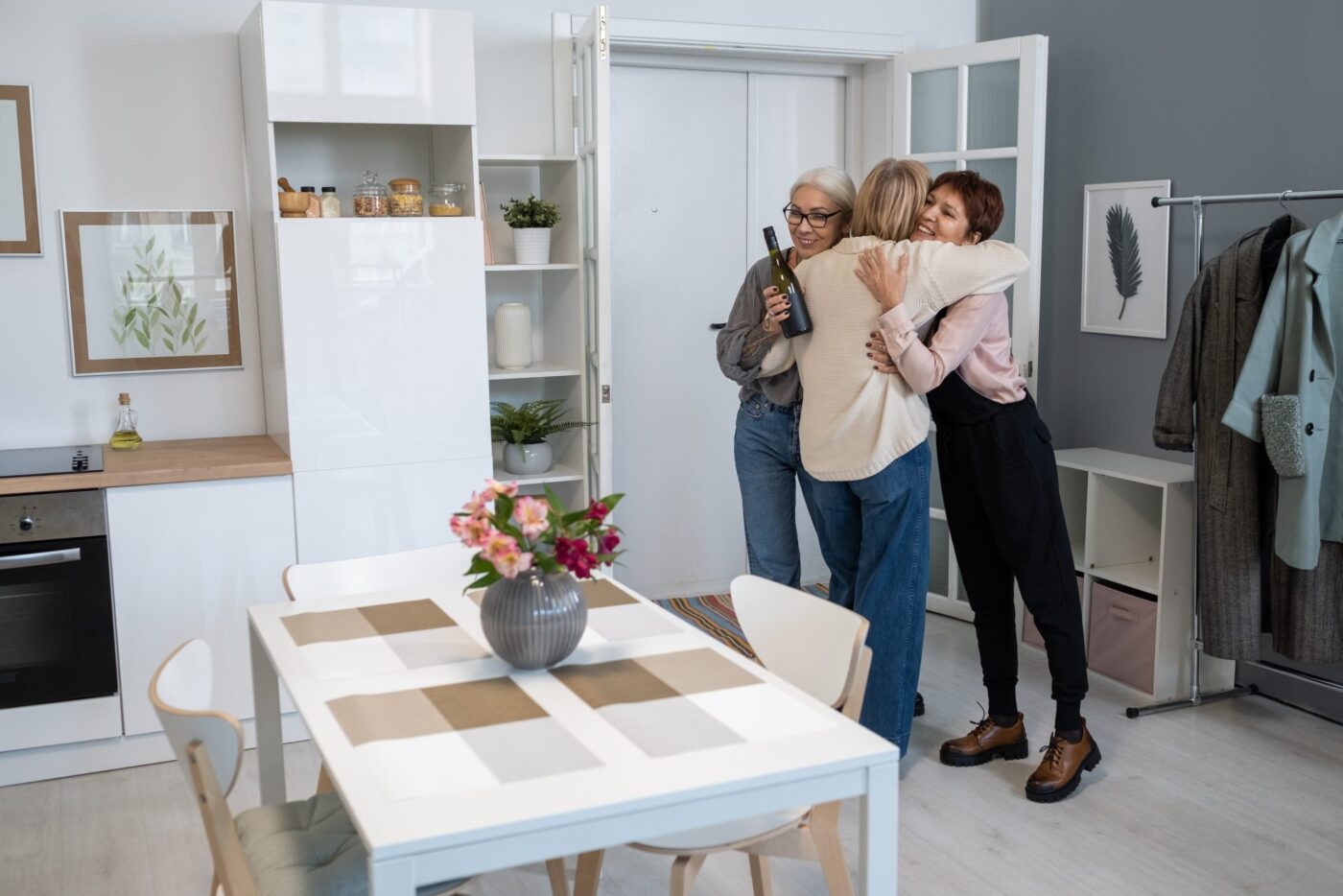 Dos mujeres abrazan a una tercera que llega con una botella de vino. Diseño de interiores, vivienda, bienes raíces, hogar.