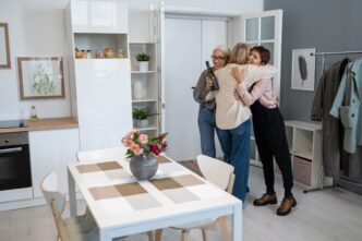 Dos mujeres abrazan a una tercera que llega con una botella de vino. Diseño de interiores, vivienda, bienes raíces, hogar.