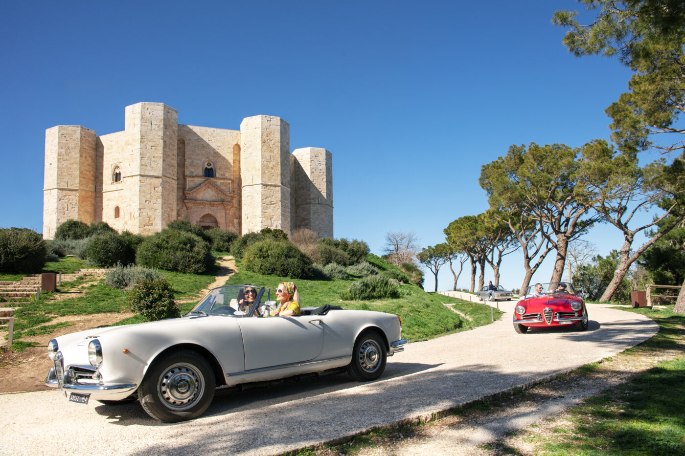 Escenario de lujo en Puglia: Dos mujeres en coche clásico frente al Castel del Monte. Cine y viajes en la costa italiana, un destino único.