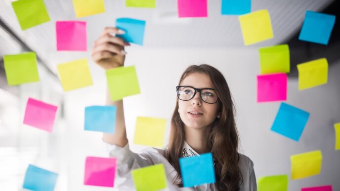 Mujer joven con gafas organizando notas adhesivas de colores en una pared de cristal. Planificación hotelera con herramienta IA Smart Summaries.
