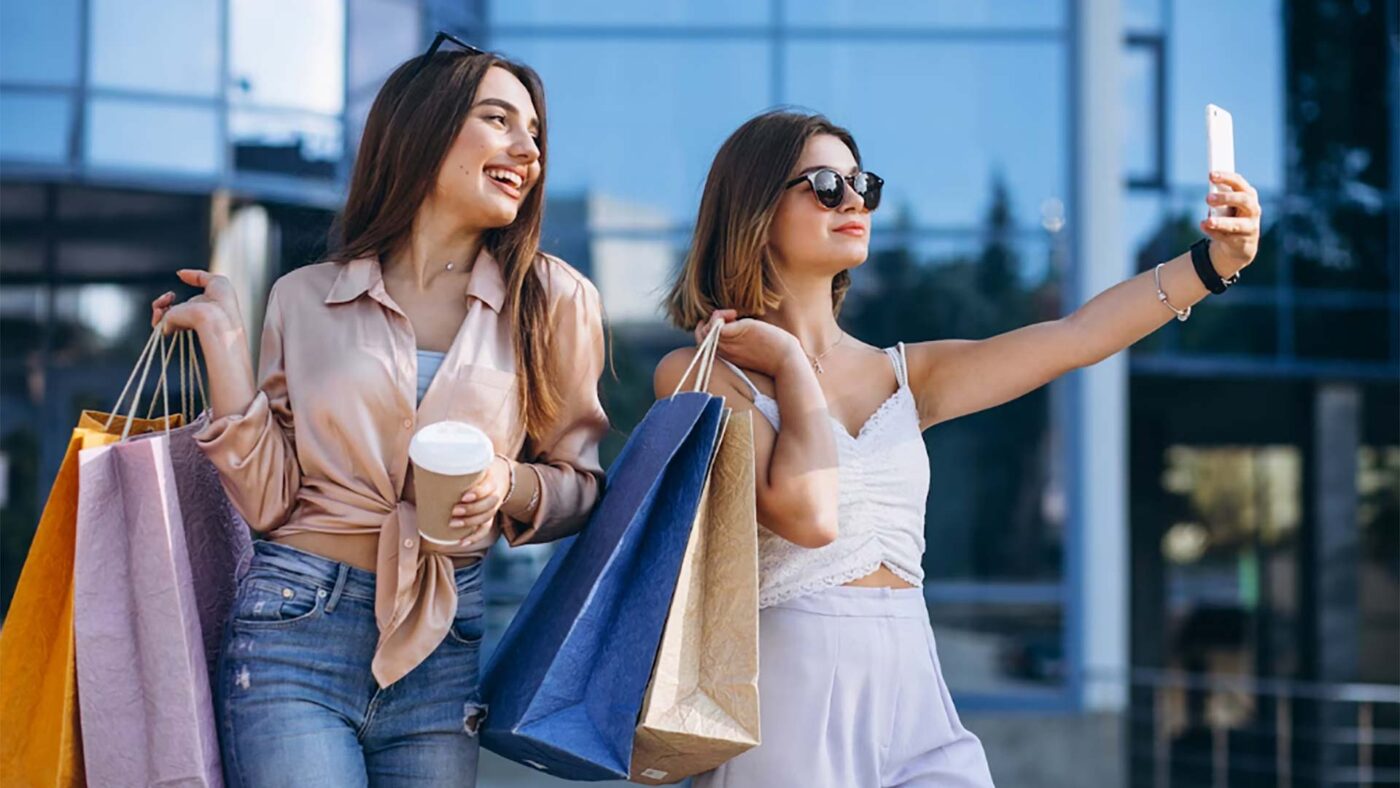 Dos amigas disfrutan del retailtainment con bolsas de compras y un café, mientras una toma una selfie en un centro comercial moderno.
