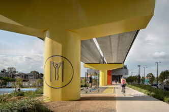 Espacio urbano revitalizado con área de calistenia bajo puente amarillo. Ciclista y persona haciendo ejercicio. Participación comunitaria activa.