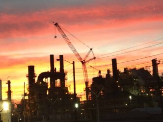 Silueta de refinería al atardecer con grúa, gestión de derechos de construcción. Cielo naranja y rosa, industria pesada. SunRay revoluciona.
