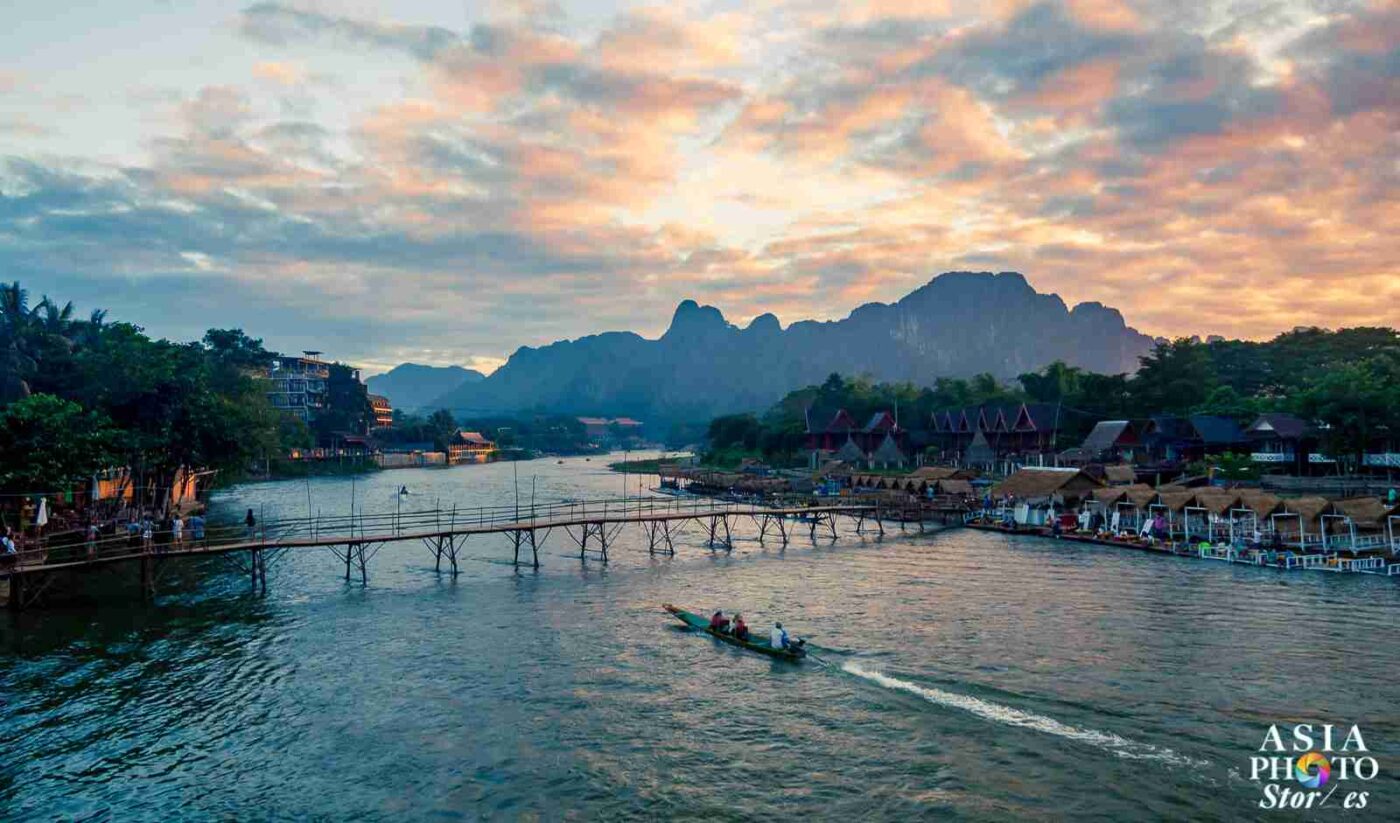Paisaje de Vang Vieng al atardecer, modelo de turismo sostenible en Asia. Río con bote, puente de madera y cabañas. Montañas al fondo.