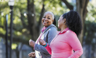 Dos mujeres afroamericanas sonrientes caminan en un parque arbolado, promoviendo la salud física y mental. Estilo de vida activo y bienestar.