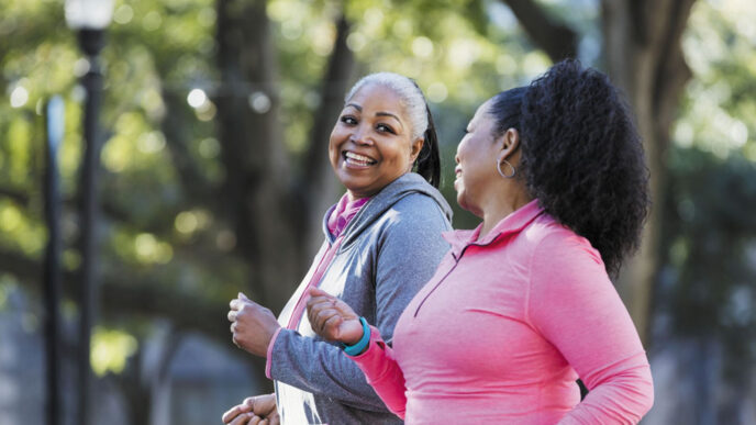 Dos mujeres afroamericanas sonrientes caminan en un parque arbolado, promoviendo la salud física y mental. Estilo de vida activo y bienestar.