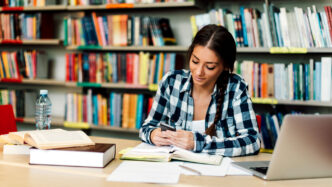 Estudiante en la Ciudad Universitaria BUAP II, 2024, usando su teléfono móvil en la biblioteca, rodeada de libros y una computadora portátil.