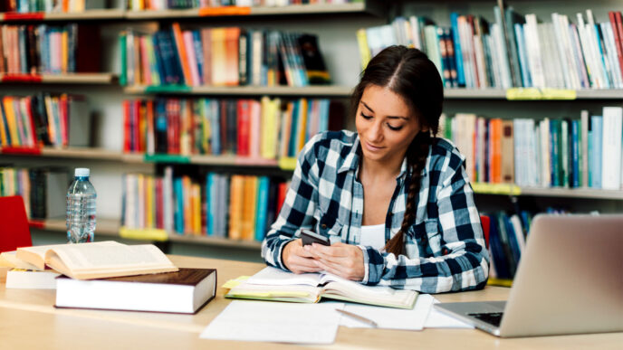Estudiante en la Ciudad Universitaria BUAP II, 2024, usando su teléfono móvil en la biblioteca, rodeada de libros y una computadora portátil.