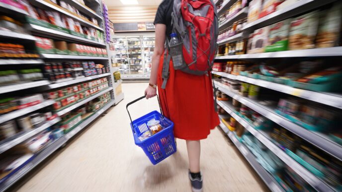 Mujer con mochila roja y cesta azul de compras en supermercado. Viajar de shopping a la baja: persona comprando en pasillo lleno de estantes.