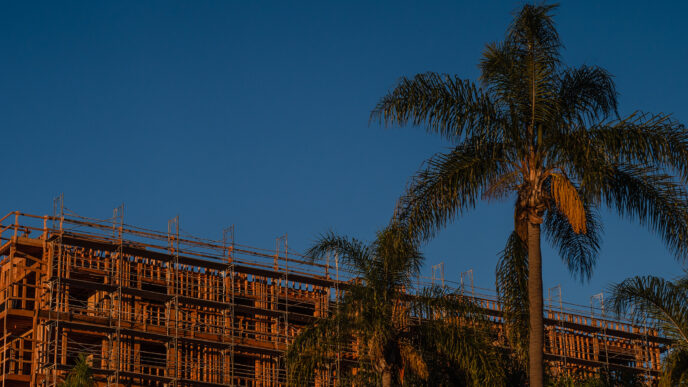 Estructura de vivienda económica en construcción, 2023. Edificio con andamios y palmeras. Caprin reporta insuficiencia de viviendas.