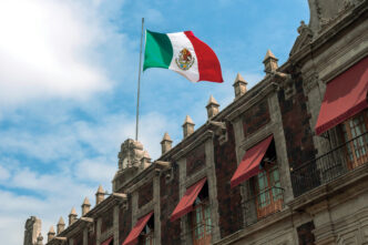 Bandera de México ondeando sobre edificio histórico. Riesgos en vivienda por criterios políticos BBVA en México. Arquitectura mexicana y cielo azul.