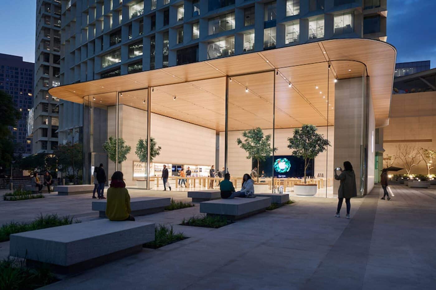Vista del Apple Store en Antara, Nuevo Polanco. Diseño moderno con paredes de cristal, árboles interiores y bancas exteriores. Iluminación cálida y gente.