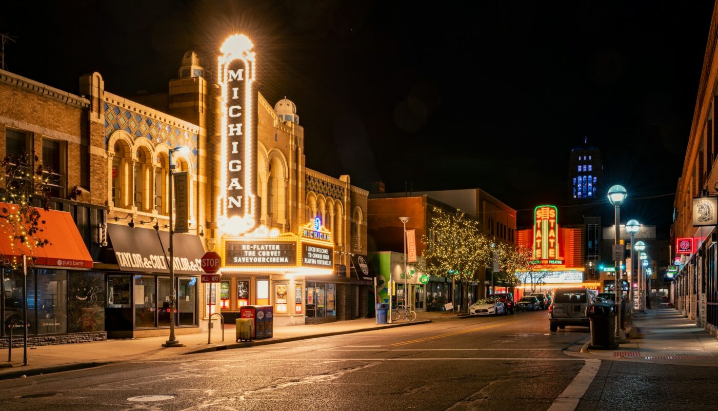 Calle del centro de noche con marquesina iluminada de Michigan Theater, tiendas y vales de vivienda visibles.