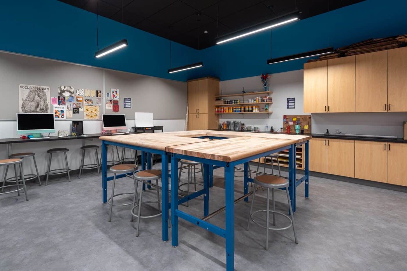 Espacio creativo con mesas azules y taburetes, computadoras iMac y estantes con materiales. Ideal para la construcción de un centro comercial.