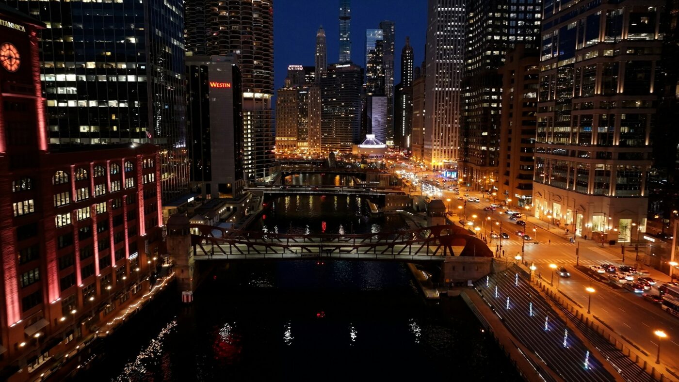 Persona cruza puente de Foundry Park sobre río, edificios iluminados y calles concurridas en centro urbano de noche.