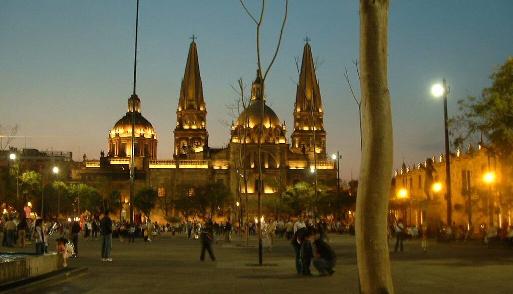 Vista nocturna de la Catedral de Guadalajara, México, con su arquitectura iluminada y gente paseando en la plaza. Destino turístico popular.