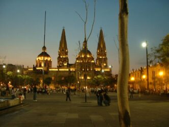 Vista nocturna de la Catedral de Guadalajara, México, con su arquitectura iluminada y gente paseando en la plaza. Destino turístico popular.