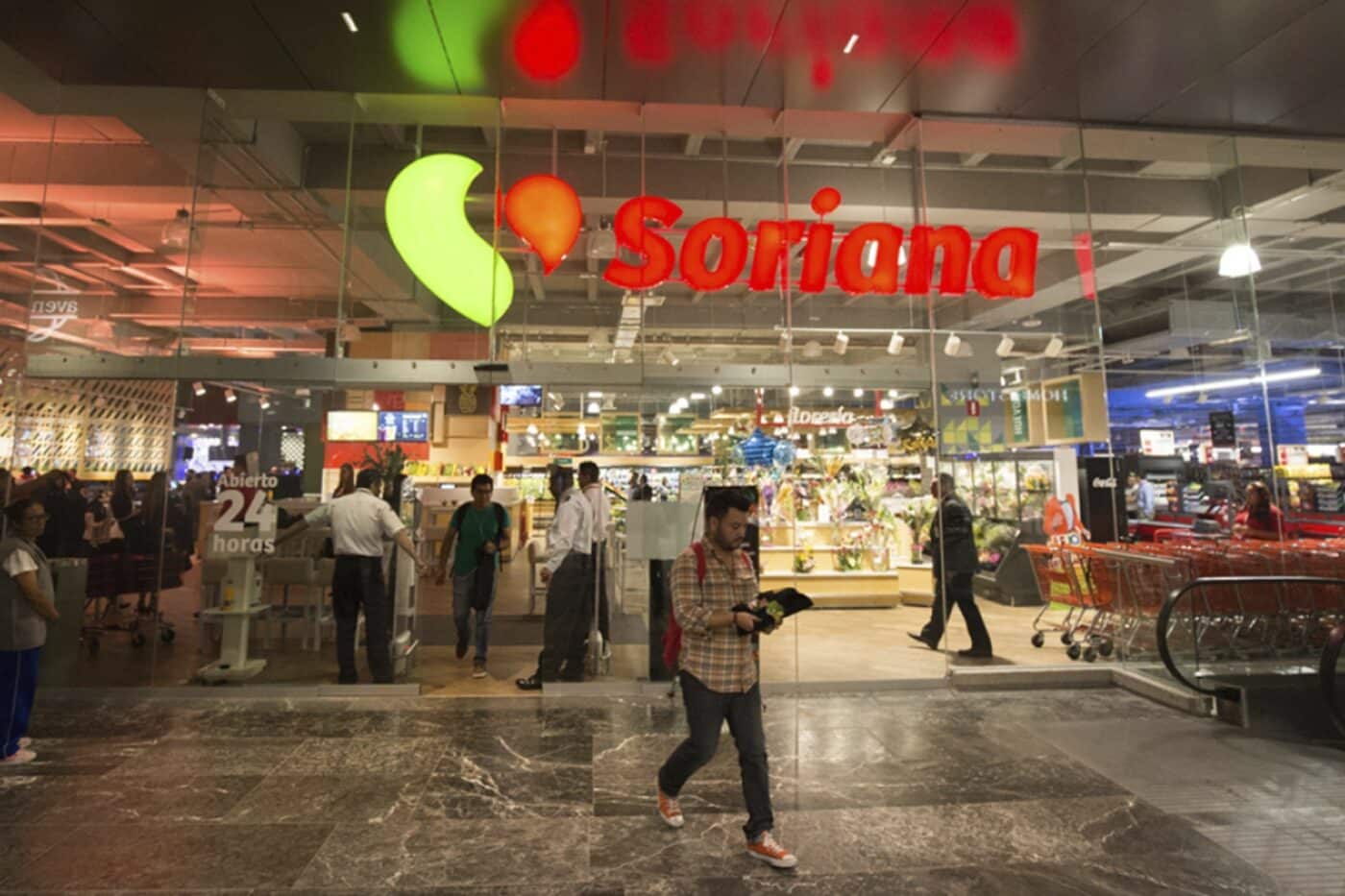 Entrada del supermercado Soriana, parte de la red de carga rápida para vehículos eléctricos en México. Gente entrando y saliendo, logo de Soriana visible.