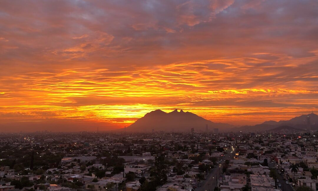 Amanecer en Monterrey, México, con el Cerro de la Silla destacando en el horizonte bajo un cielo naranja y nubes espectaculares. Vista panorámica.