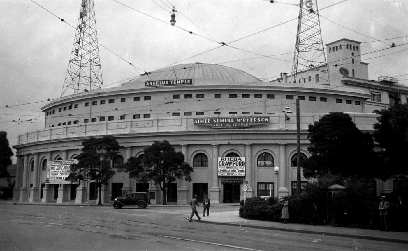 Vista del Angelus Temple, con su arquitectura distintiva y torre de radio. La imagen muestra la entrada principal con letreros y personas.