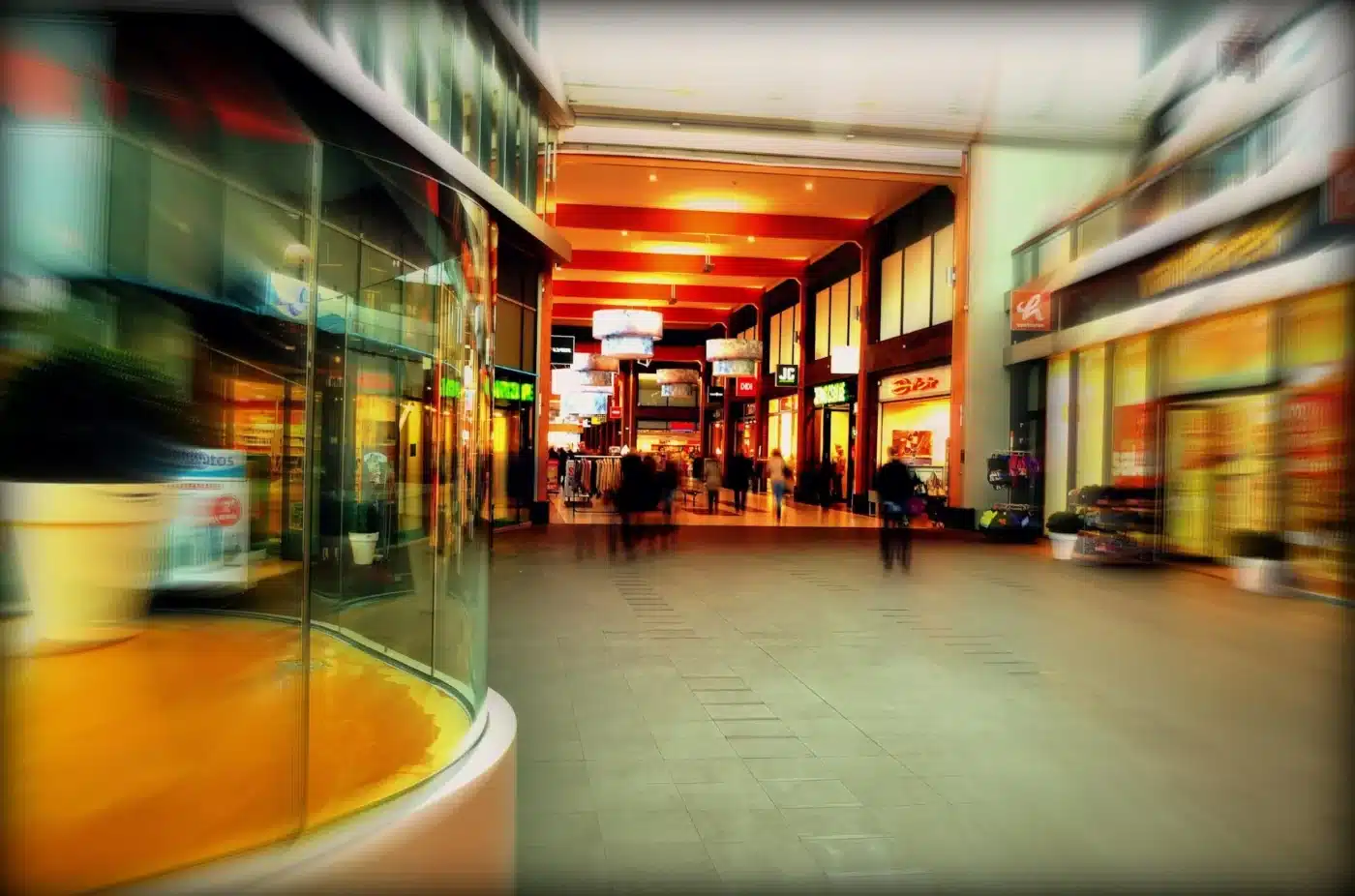 Personas caminando en pasillo de centro comercial, tiendas Sak's Global y otros locales al fondo.