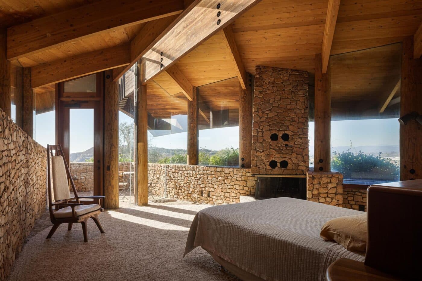 Persona en recámara con muros de piedra y madera, techo alto y luz natural, vista a huerto de aguacates.