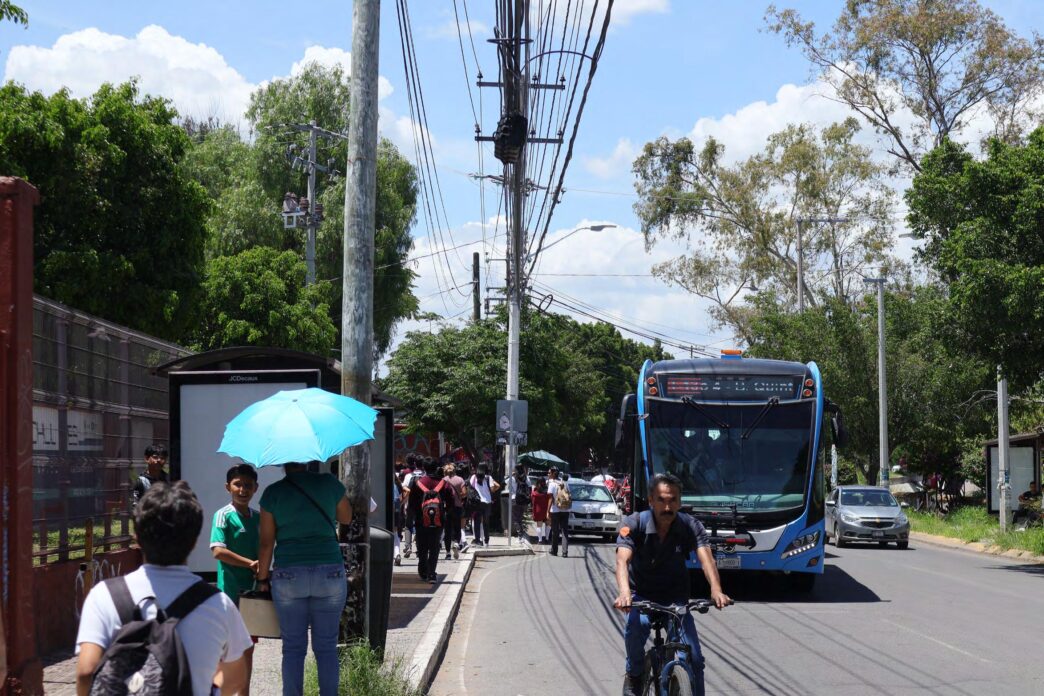 Escena urbana en Culiacán: autobús de transporte público moderno, ciclista y personas esperando bus. Movilidad sostenible y acceso en primer plano.