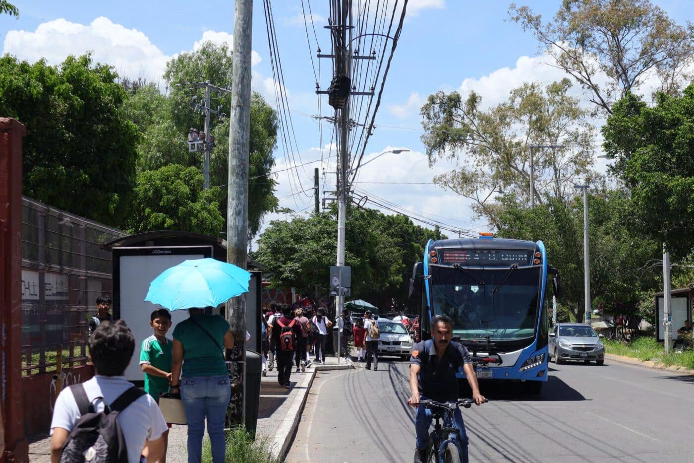 Escena urbana en Culiacán: autobús de transporte público moderno, ciclista y personas esperando bus. Movilidad sostenible y acceso en primer plano.